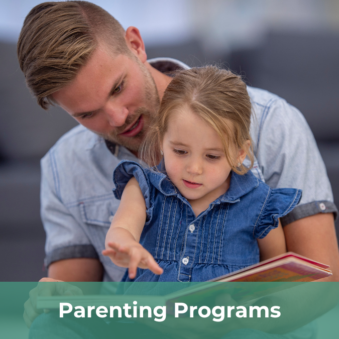 A young man sits with his daughter, reading a book to her. There is a text bar on the bottom of the image that reads Parenting Programs.